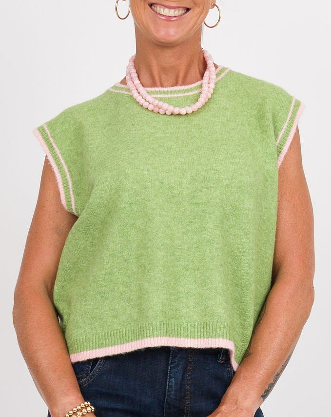 A woman wearing a sleeveless knit sweater vest in light green with pale pink trim, paired with dark blue jeans and a pink beaded necklace, against a plain white background.