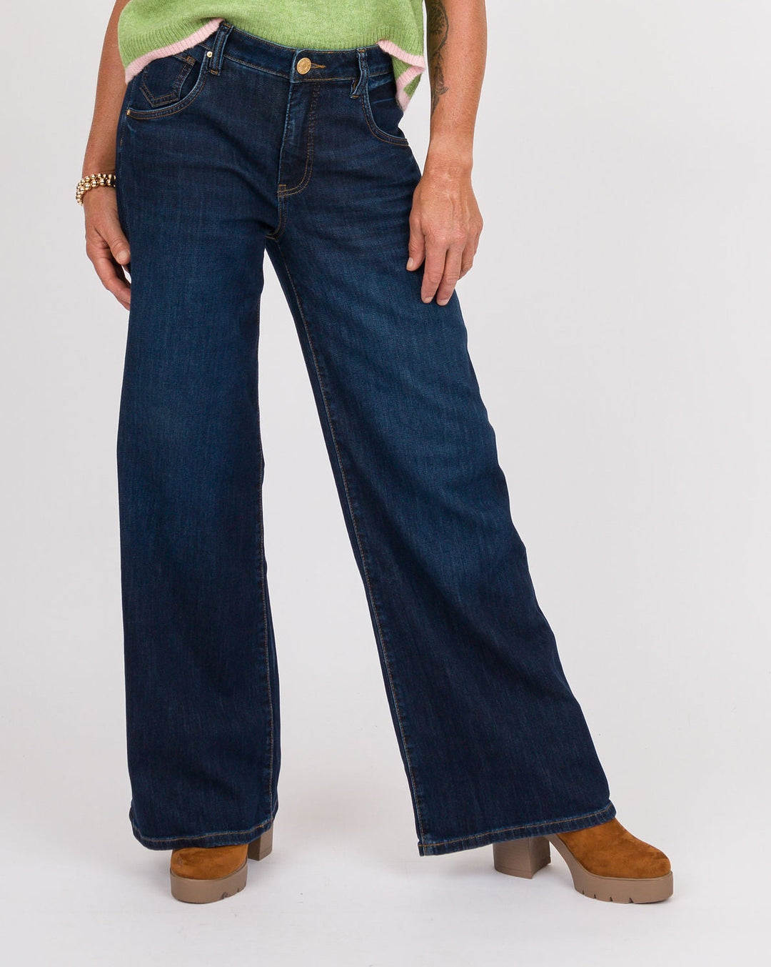 A woman modeling a light green sleeveless knit sweater vest with cream trim, paired with dark blue wide-leg jeans and brown shoes, against a plain white studio background.