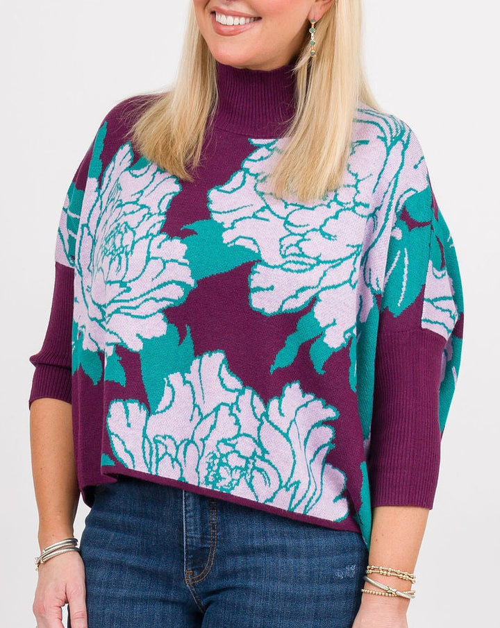 A woman modeling a color-block knit mock-neck sweater with a floral pattern in teal, white, and deep purple, paired with blue jeans, brown clogs, and holding a matching purple clutch against a white studio background.