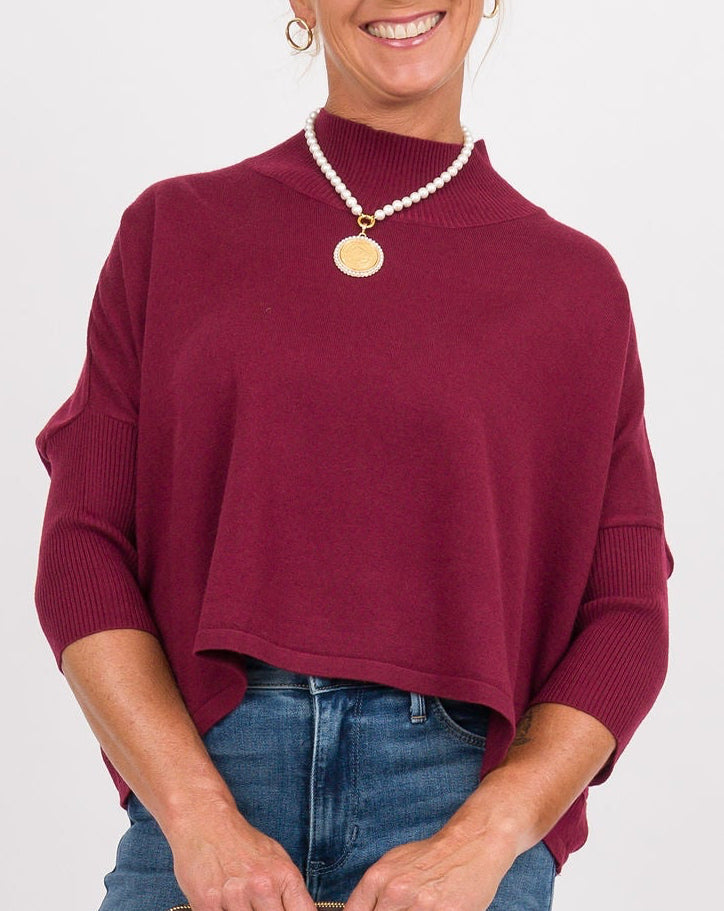A woman modeling a dark red long-sleeve knit top and blue flared jeans, holding a small dark clutch against a plain white studio background.