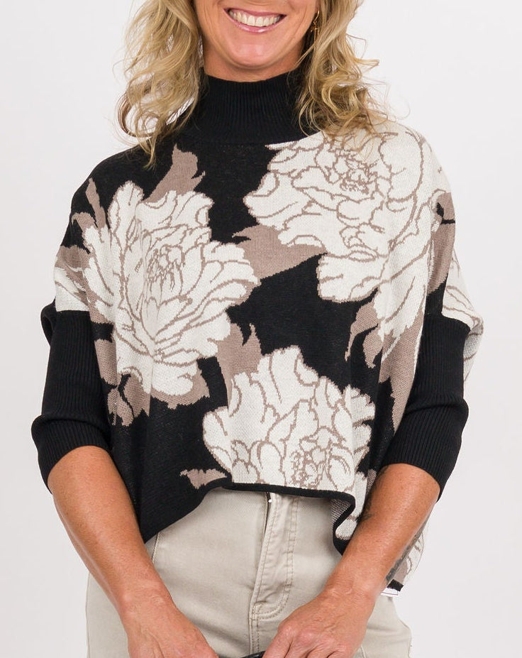 A woman modeling a black long-sleeve turtleneck top with a large beige and white floral print, paired with light beige wide-leg pants, a black woven clutch, and clear high-heeled sandals, against a plain white studio background.