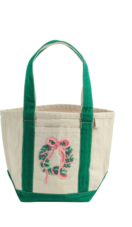 A canvas tote bag with green handles and bottom trim, featuring a pink and green embroidered wreath with a bow on the front pocket, set against a plain white background.