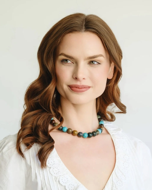 A woman with wavy brown hair wearing a colorful beaded necklace and a white top, photographed against a plain light background.