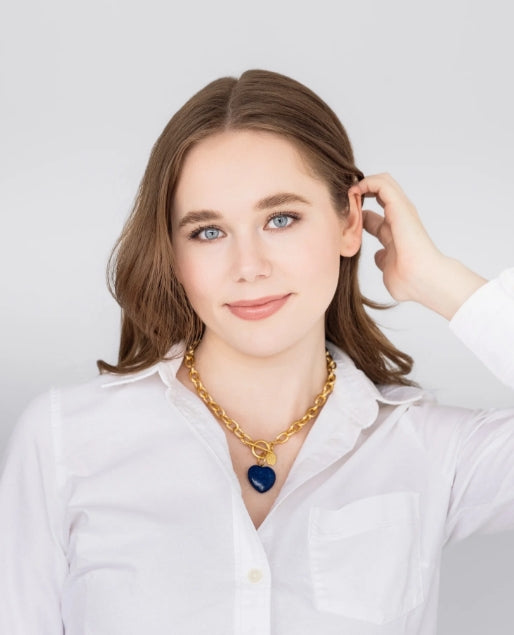 A woman with brown hair wearing a white button-up shirt and a gold chain necklace with a dark blue heart-shaped pendant, posing against a light gray background.