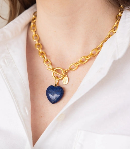 A close-up of a gold chain necklace with a toggle clasp and a polished dark blue heart-shaped pendant, worn by a person in a white collared shirt.