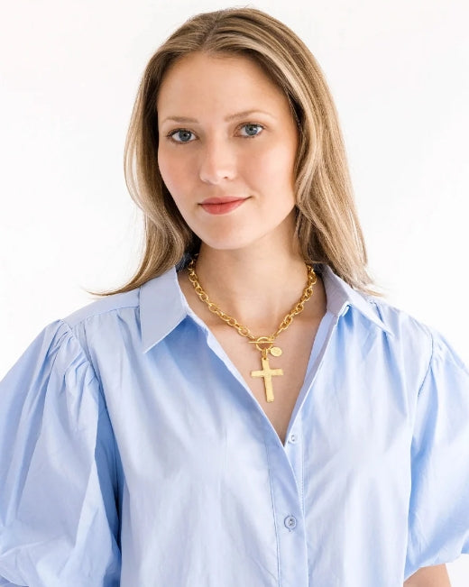 A woman with blonde hair wearing a light blue button-up shirt and a gold chain necklace with a cross pendant, photographed against a plain white background.