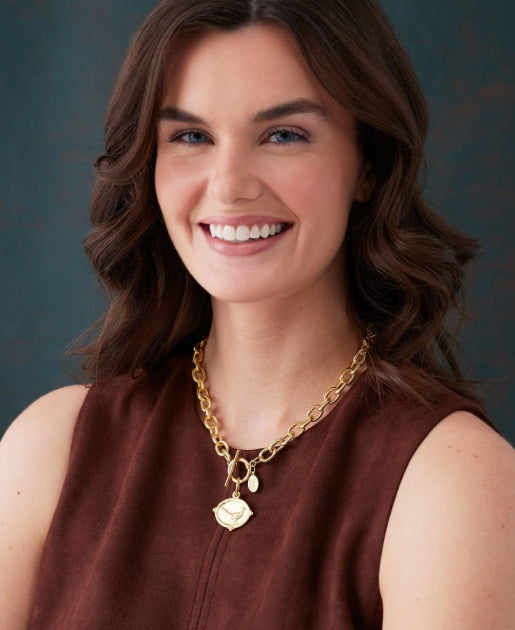 A smiling woman with brown hair wearing a gold chain necklace with a small heart-shaped charm, paired with a brown sleeveless top, set against a dark textured background.