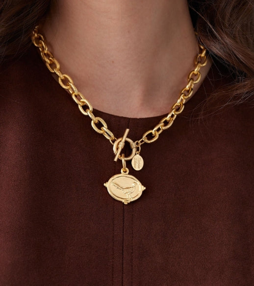 A close-up of a gold chain necklace with a toggle clasp and two charms, worn by a person in a brown top. One charm is a lemon-shaped pendant with a bird design, and the other is a small oval tag.