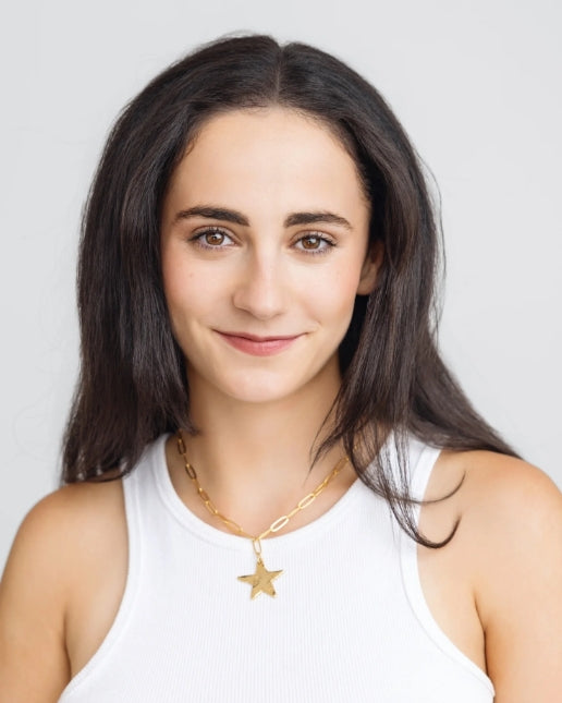 A woman with long dark brown hair wearing a white ribbed tank top and a gold star pendant necklace, photographed against a light gray background.