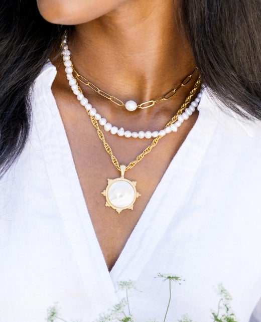 A close-up of a model wearing three layered necklaces: a white pearl strand, a gold paperclip chain with a pearl, and a gold chain with a sunburst pendant featuring a white circular center.