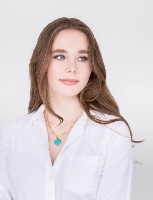 A young woman with long brown hair wearing a white button-up shirt and a gold necklace with a turquoise heart pendant, set against a plain white background.