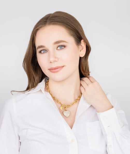 A woman with light brown hair wearing a white button-down shirt and a gold chain necklace with a heart-shaped pendant, posing against a light gray background.