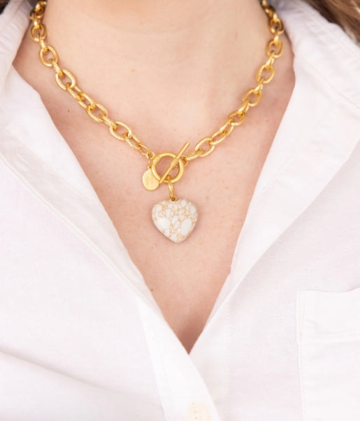 A close-up of a gold chain necklace with a toggle clasp and a heart-shaped stone pendant, worn by a person in a white collared shirt.