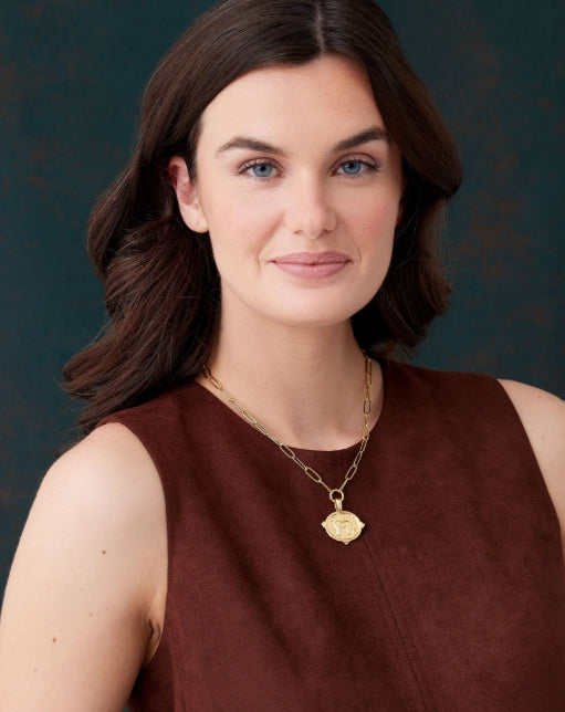 A woman with dark brown hair and blue eyes wearing a brown sleeveless top and a gold pendant necklace, photographed against a dark textured background.
