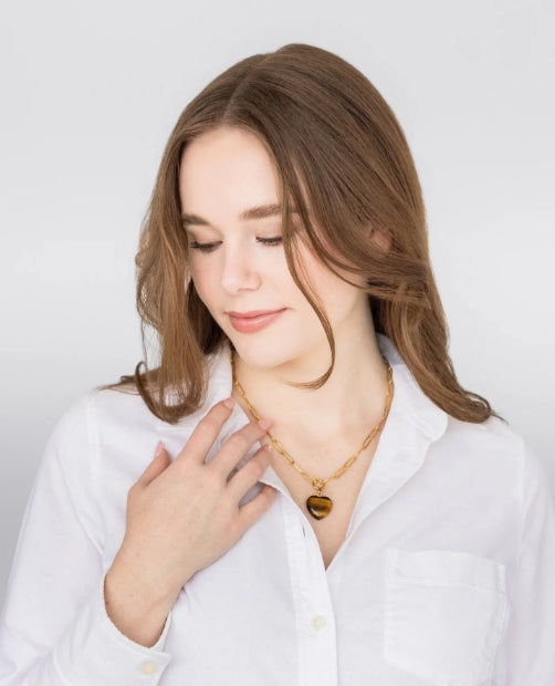 A woman with light brown hair wearing a white button-up shirt and a gold chain necklace with a heart-shaped pendant, posing against a light gray background.