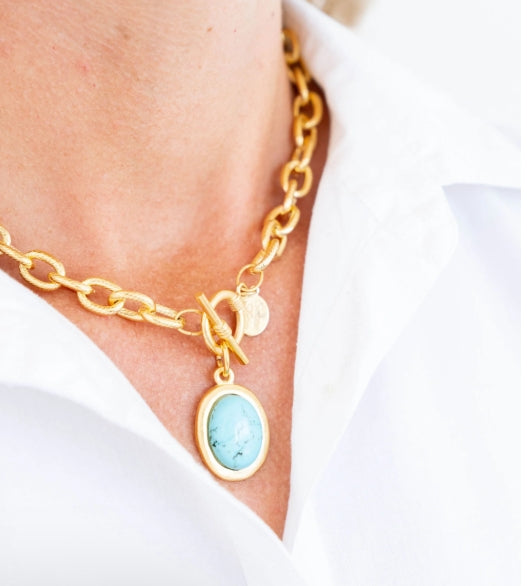 A close-up of a gold chain necklace worn by a person, featuring a toggle clasp and an oval pendant with a light blue stone in a gold bezel, set against a white collared shirt.