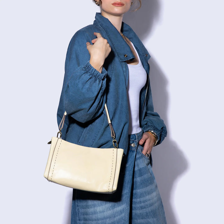 A woman wearing a blue denim jacket and matching wide-leg jeans carries a cream-colored leather shoulder bag with gold-tone hardware against a light gray studio background.
