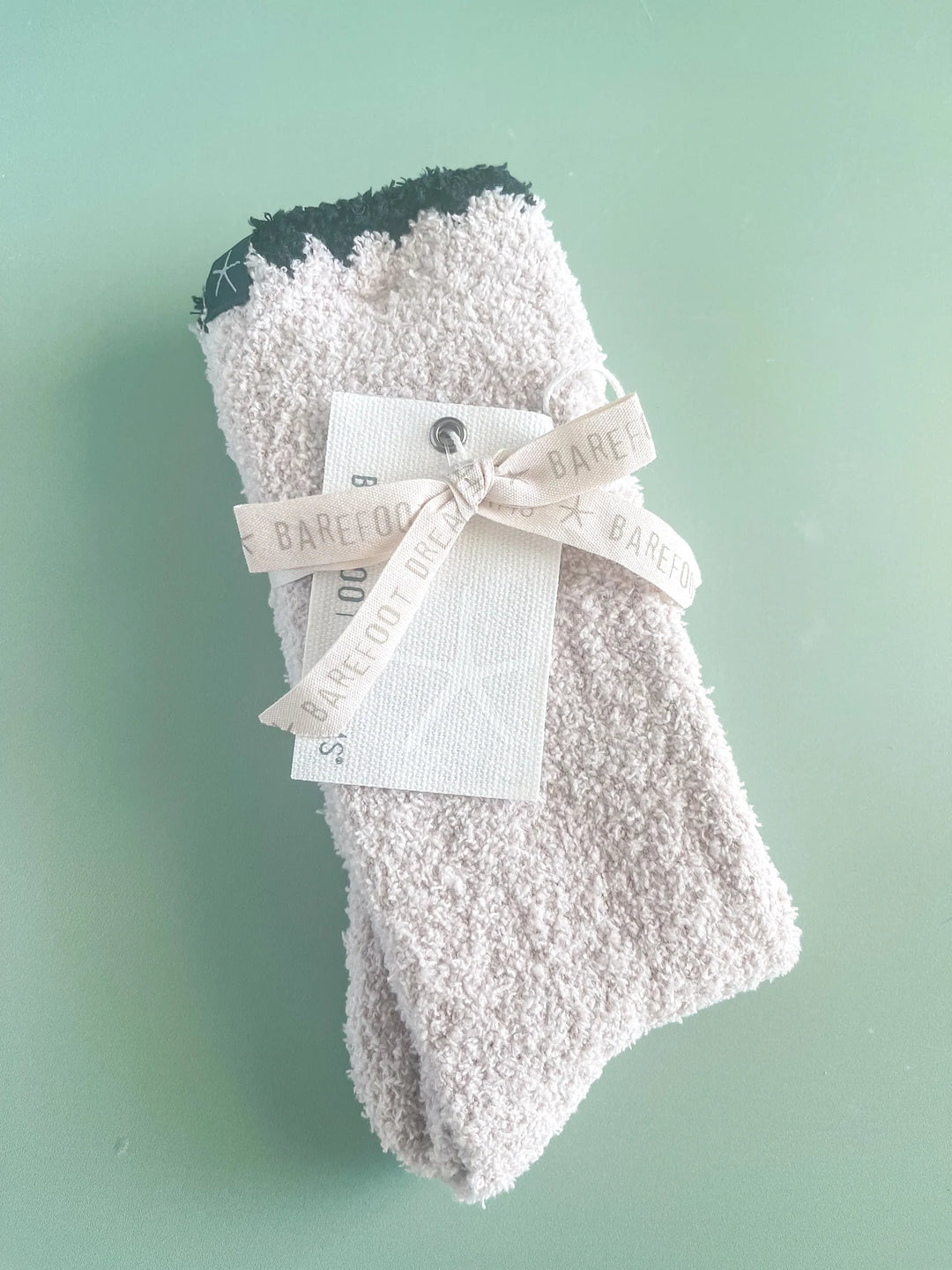 A pair of light beige terry cloth socks with a black toe cap, tied with a cream ribbon and tag, on a mint green background.