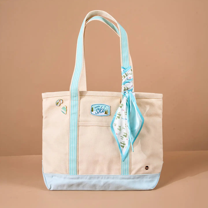 A beige canvas tote bag with light blue striped handles, a light blue bottom trim, and a matching light blue patterned scarf tied around one handle. The bag has a front pocket with a blue brand patch reading &
