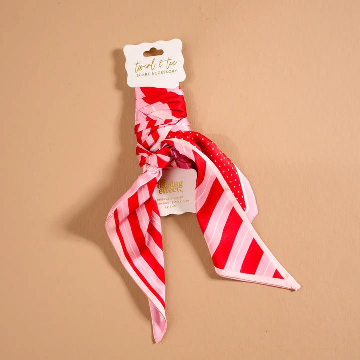 A red and pink striped scarf accessory in its original white cardboard packaging, displayed against a solid beige background.