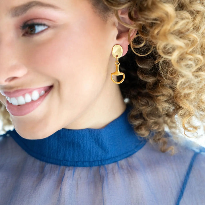A close-up of a smiling woman with curly blonde hair wearing a gold dangle earring with a small oval stud and a teardrop-shaped drop, paired with a blue high-neck sheer top.