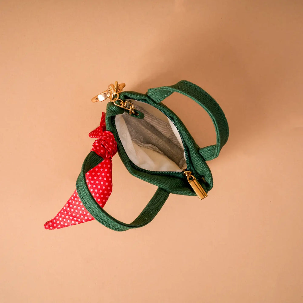 A small green canvas zippered pouch with a red and white polka dot scarf tied to the zipper, shown on a solid beige background.
