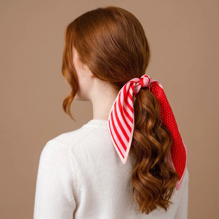 A woman with wavy red hair is shown from the back, wearing a white knit sweater and a red and white striped scarf tied in her ponytail against a solid beige background.