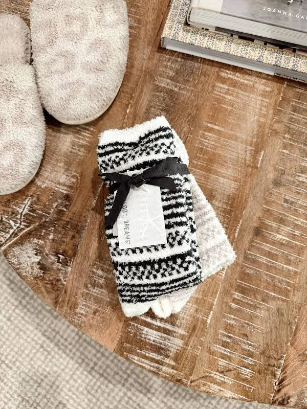 A pair of knit toe socks with a black and white geometric pattern, tied with a black ribbon and a white paper tag, displayed on a rustic wooden table next to a pair of beige slippers.